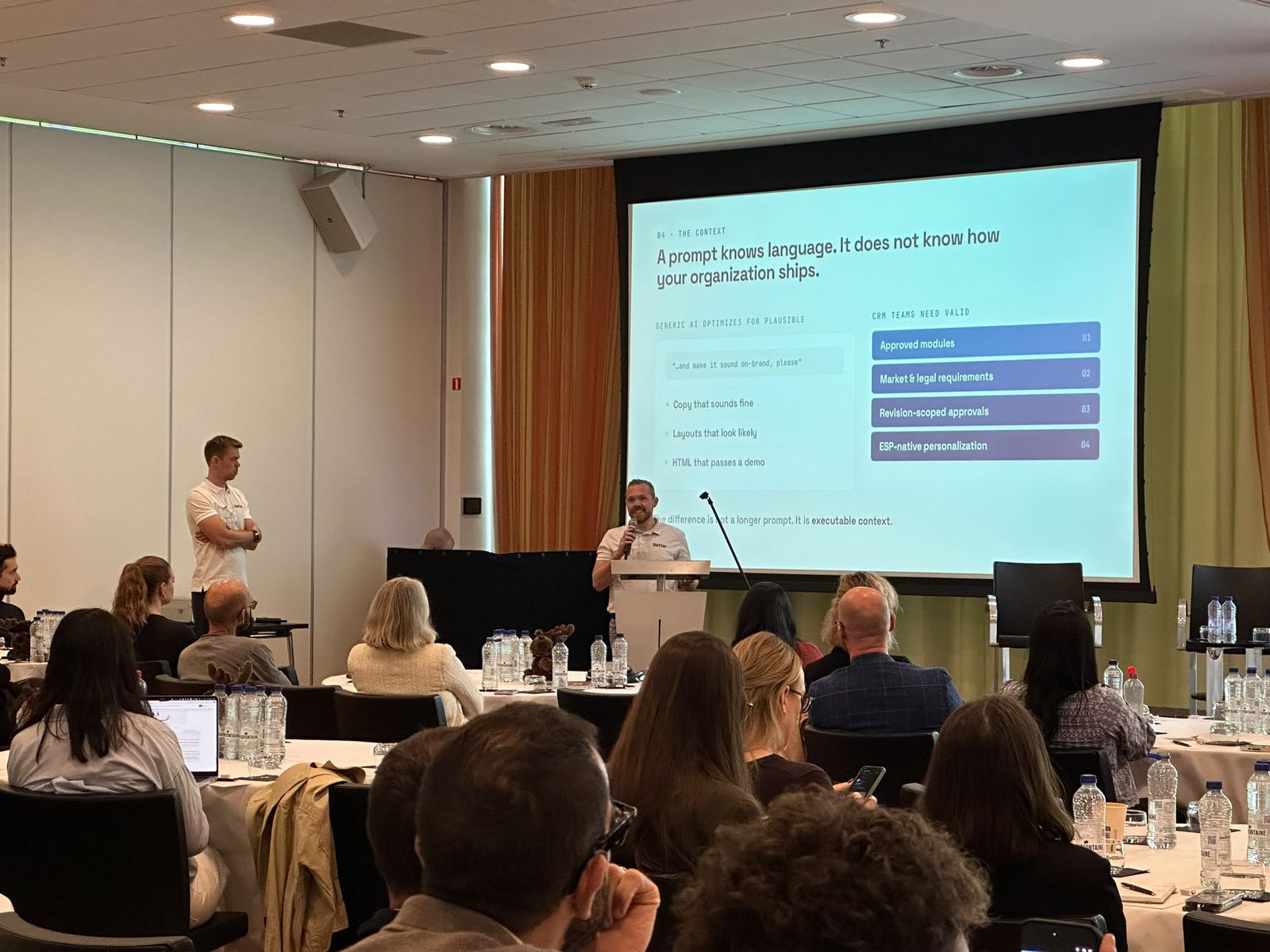 Nicki presenting the slide "A prompt knows language. It does not know how your organization ships." to the audience in Amsterdam, with Simon standing to the side
